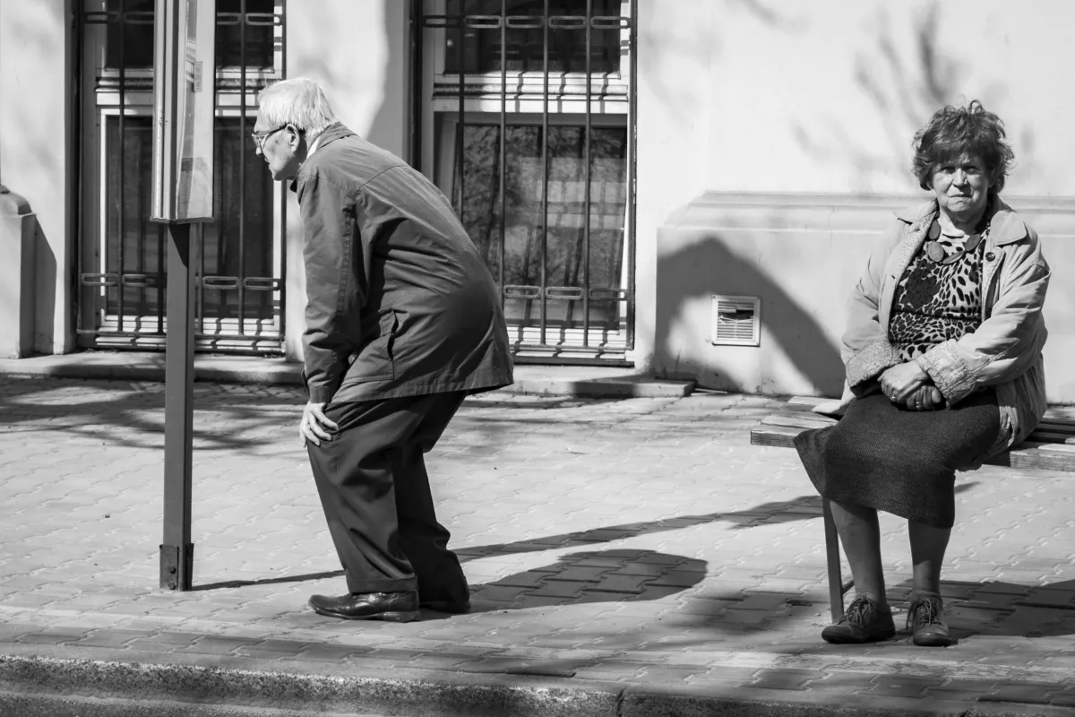 Older man preparing to cross while a woman sits on a chair at the curb